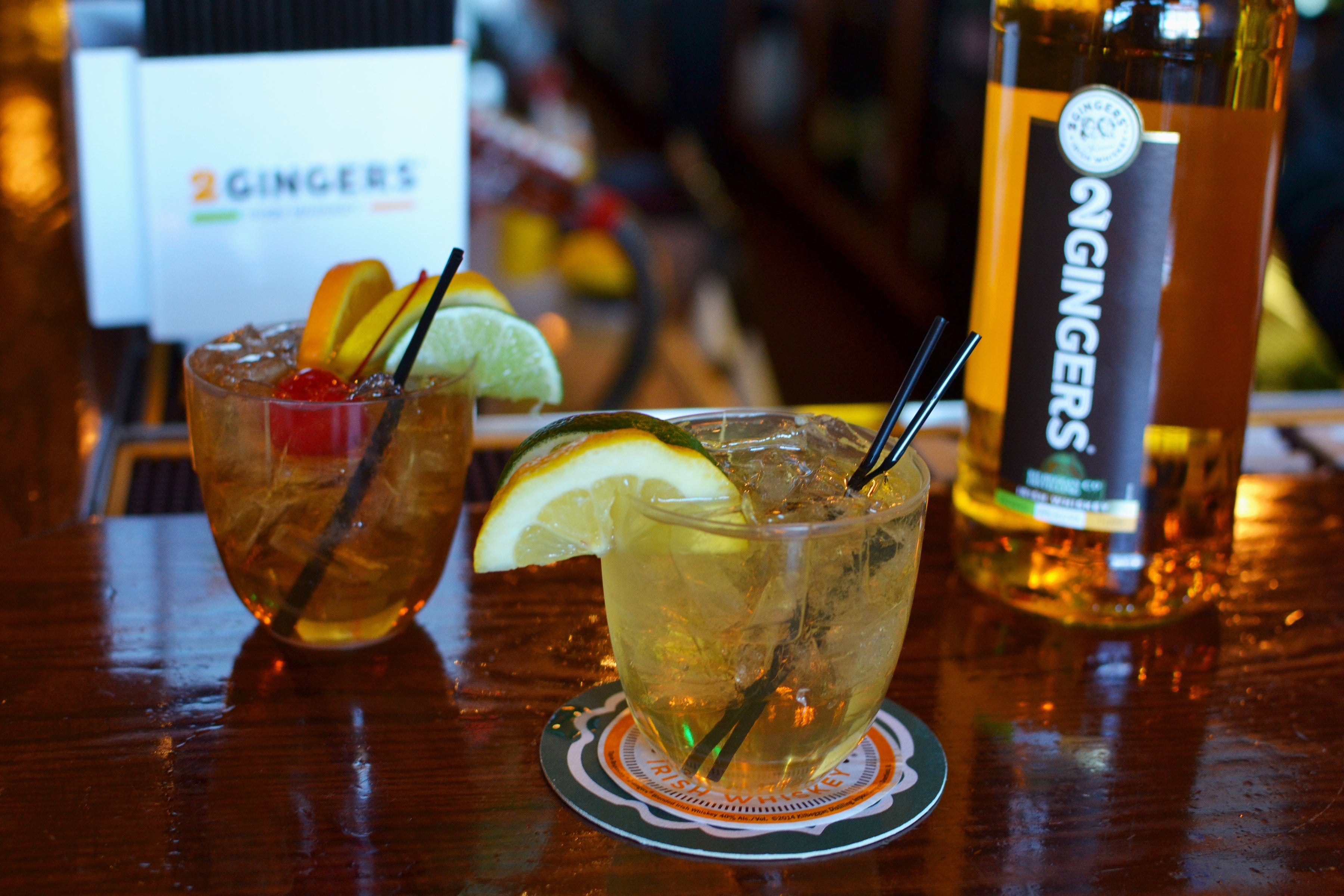 Compostable drinkware including low ball tumblers and martini-style cups filled with colorful cocktails, garnished with citrus slices and edible flowers, displayed on a wooden bar surface.
