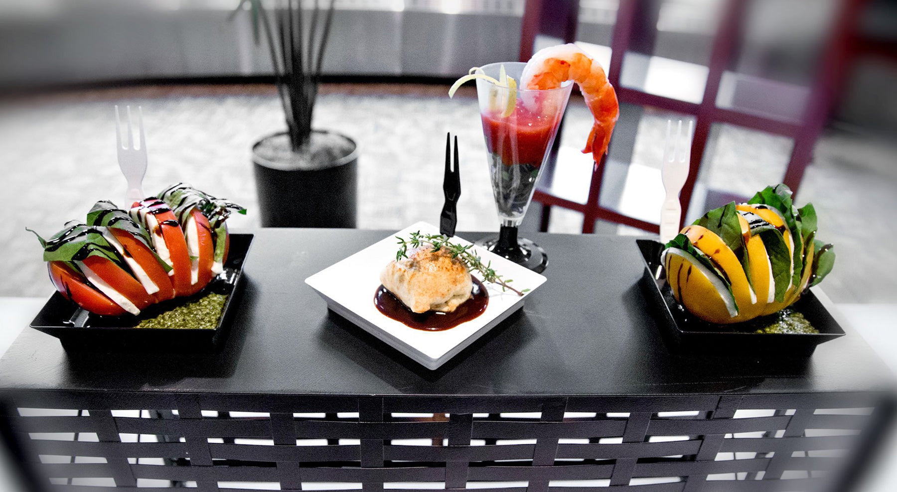 Assorted gourmet appetizers served in compostable foodware, including tomato caprese salads, puff pastry with sauce, and shrimp cocktail in a clear PLA flute, displayed on a black serving table.